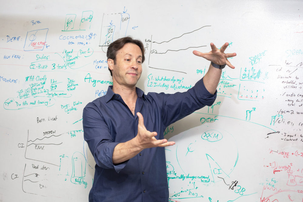 A photo of David Eagleman making hand gestures in front of a whiteboard covered in writing